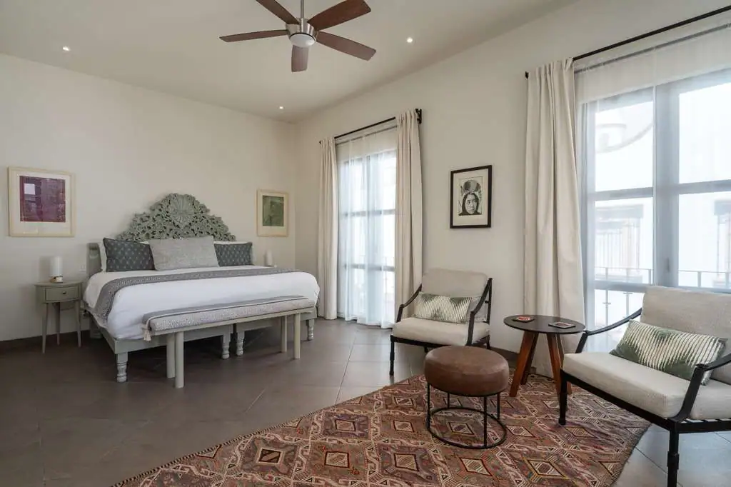 Bright hotel-style bedroom with a large ornate headboard, king bed, nightstands, and neutral decor. Large windows with white curtains illuminate the room, San Miguel de Allende