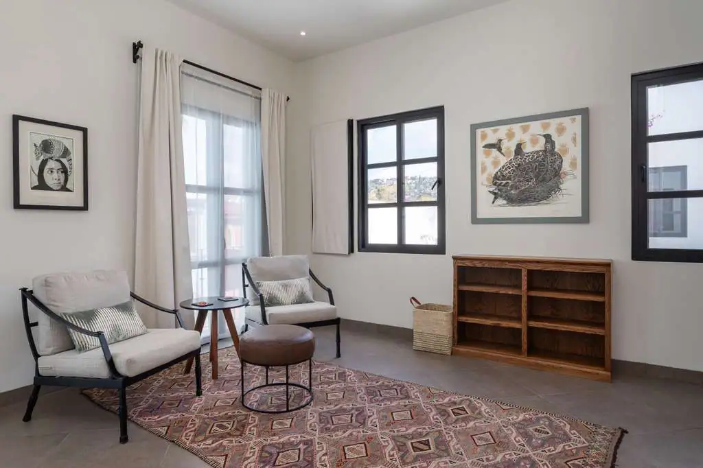 Cozy living room with two cream upholstered chairs, a small round table, and a patterned rug near a large window panelled with white curtains, San Miguel de Allende
