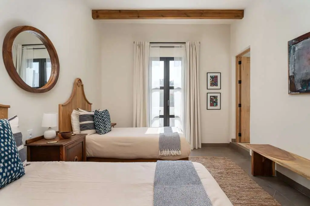 Hotel room with two twin beds, wooden headboards, round wall mirror, nightstand, lamp, and blue patterned pillows. Large window with white curtains, San Miguel de Allende
