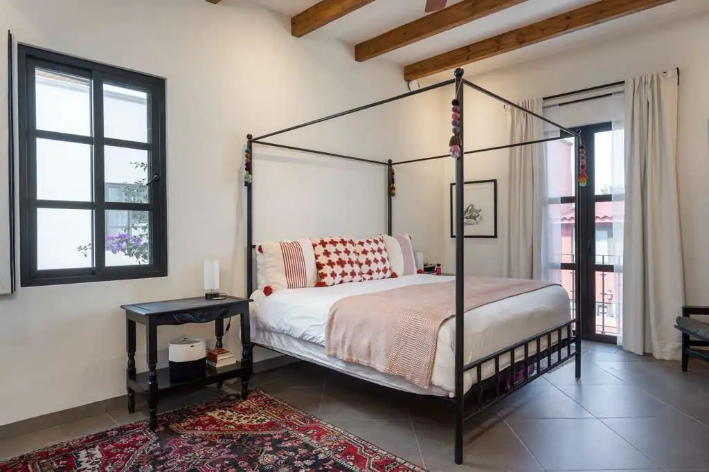 Modern black metal four-poster bed in a bright bedroom with white walls, exposed wooden beams, and a patterned rug beneath, San Miguel de Allende