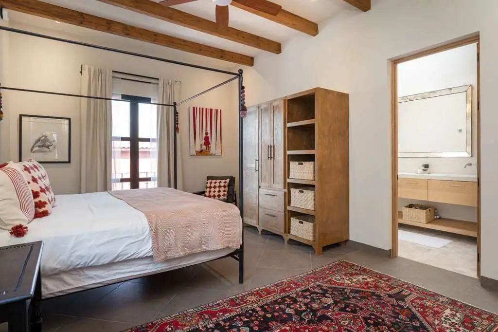 A bright bedroom with a black metal four-poster bed, white linens, red patterned pillows, and a red patterned rug; wooden ceiling beams and a large wooden wardrobe with baskets nearby. An open doorway reveals a modern bathroom, San Miguel de Allende