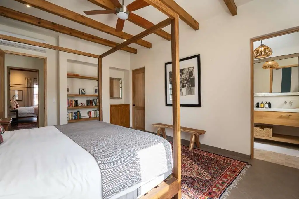 Bedroom with a wooden four-poster bed, woven rug, and built-in shelves along the wall; doorway to an en-suite bathroom visible to the right, San Miguel de Allende