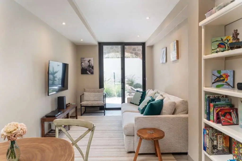 Cozy living room with beige walls, a wall-mounted TV, and a light sofa with teal cushions facing a glass door to the garden, San Miguel de Allende