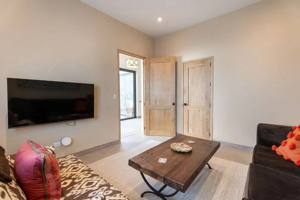 Living room with a wall-mounted TV, wooden doors, and a wooden coffee table between a patterned sofa and a dark couch with orange pillows, San Miguel de Allende