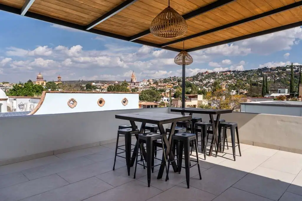 Rooftop terrace with a tall rectangular table and black stools, wicker pendant lights overhead, and a city skyline of hills and domed buildings in the distance, San Miguel de Allende