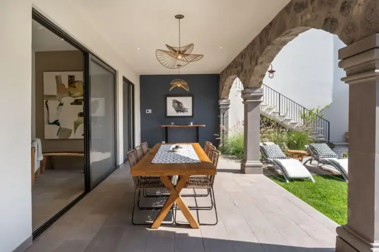 Outdoor patio with a wooden dining table and six wicker chairs under stone arches; lounge chairs and greenery beyond, San Miguel de Allende