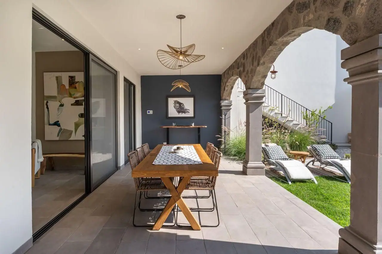 Outdoor patio with a wooden dining table and six wicker chairs under stone arches; lounge chairs and greenery beyond, San Miguel de Allende