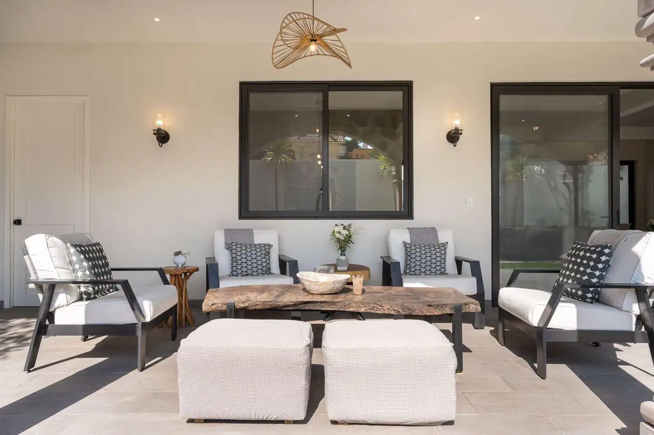 Modern outdoor patio seating: six white cushioned chairs and two matching ottomans around a rustic wooden coffee table, with decorative pillows and a small vase centerpiece under a pendant light, San Miguel de Allende