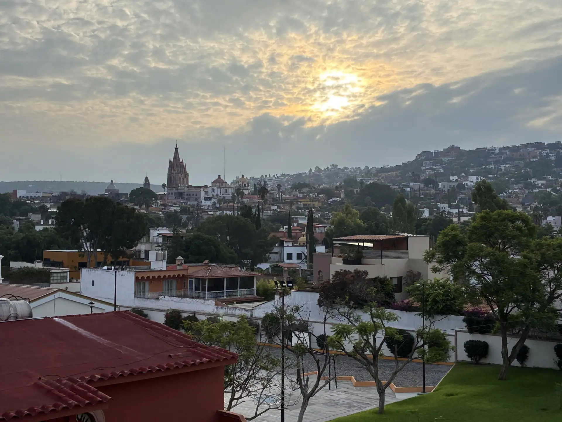 Hillside cityscape with a tall, spired church center-left, red-tiled roofs in the foreground, and a cloudy sky above, San Miguel de Allende