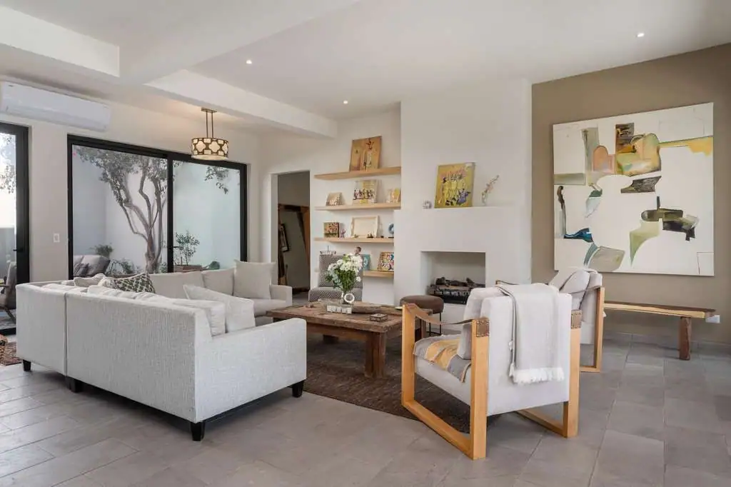 Bright modern living room with a gray sectional, wooden armchairs, a coffee table, built-in shelves, a fireplace, abstract wall art, and large glass doors opening to a patio, San Miguel de Allende