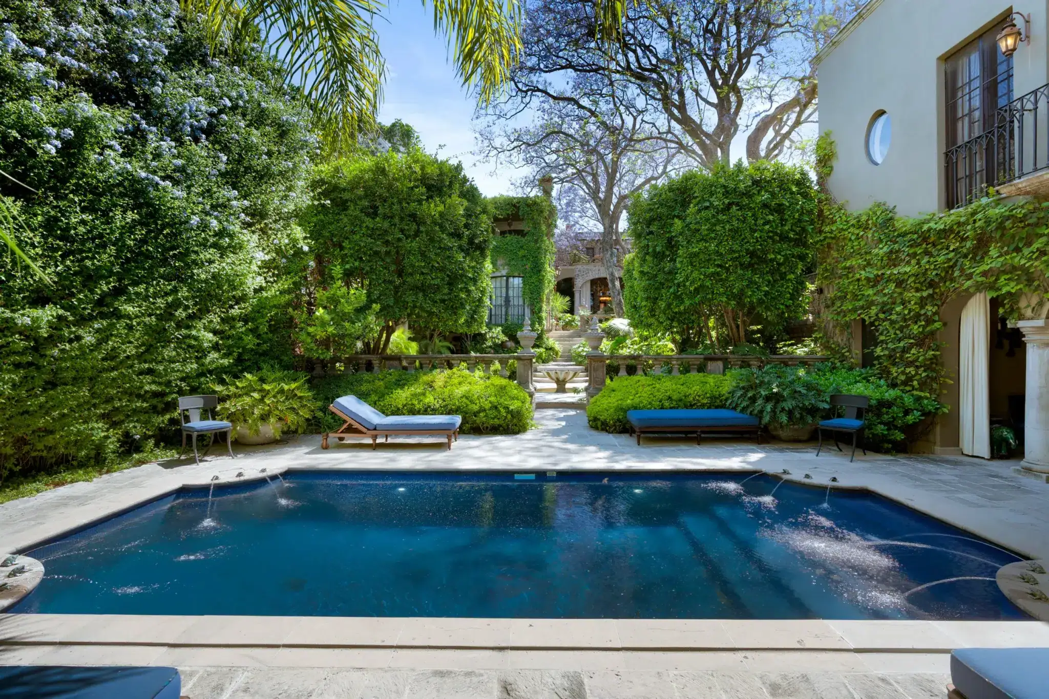 Stone pool surrounded by lush greenery and ivy-covered walls, with blue lounge chairs along the pool deck in a sunny courtyard villa setting, San Miguel de Allende