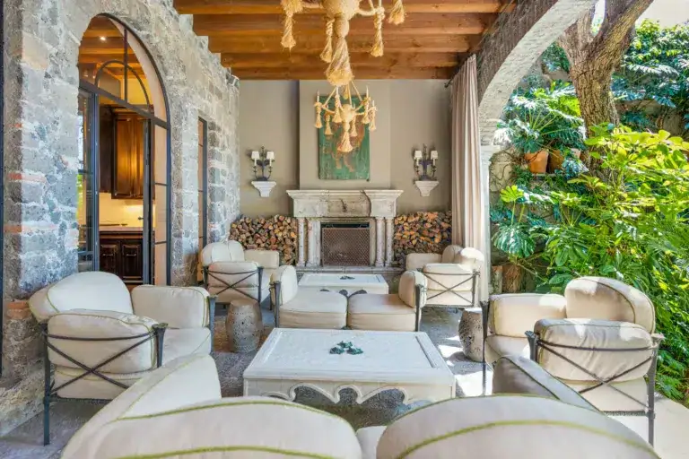 Stone-walled outdoor living room with cream cushioned chairs around a white coffee table, and a stone fireplace with stacked firewood, San Miguel de Allende