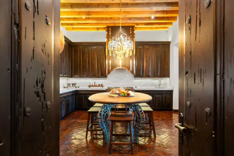 Rustic kitchen with dark wood cabinets, a crystal chandelier, and a round wooden table topped with fruit centerpiece viewed through heavy wooden doors, San Miguel de Allende
