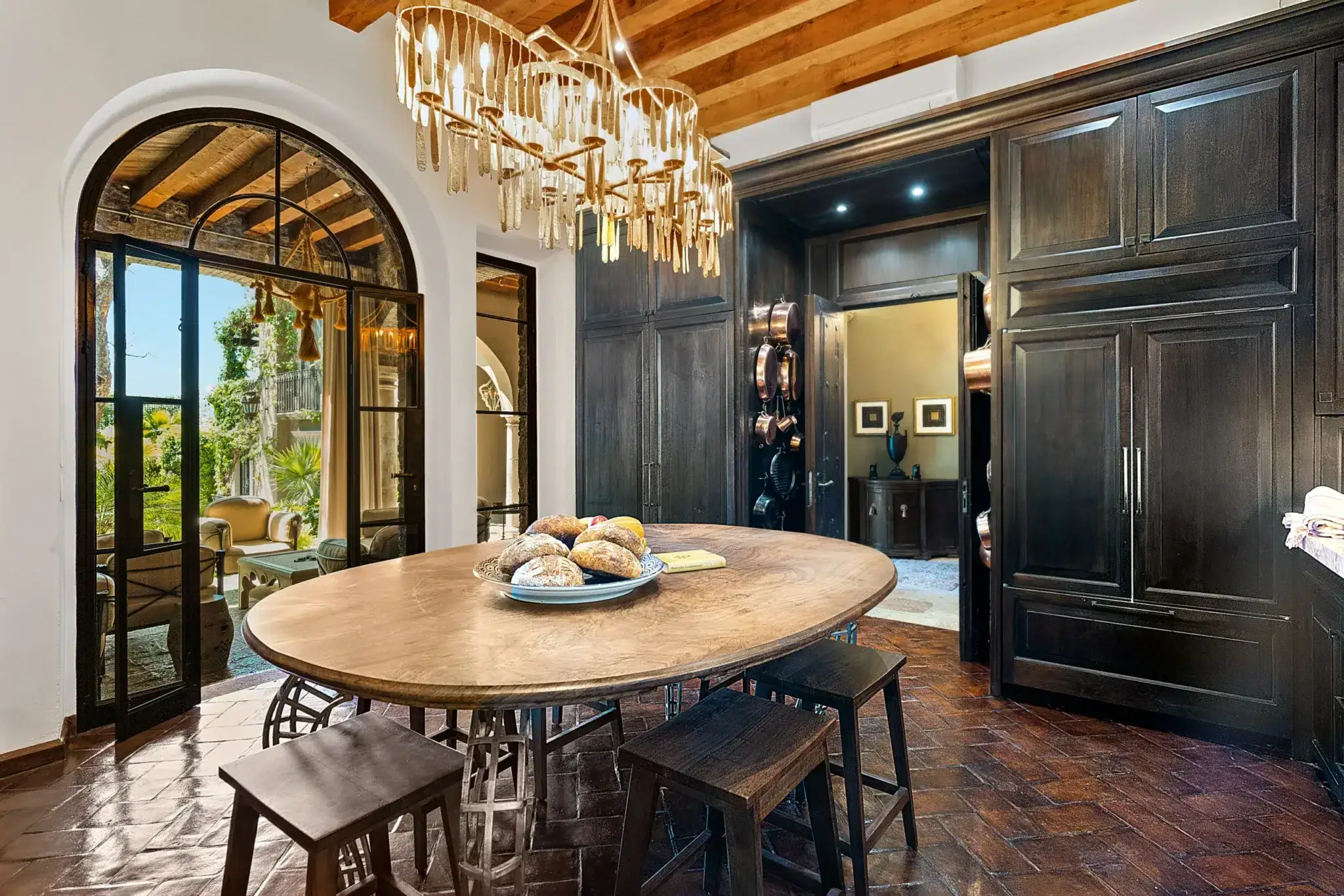 Round wooden dining table with a platter of bread in a dark wood kitchen featuring arched glass doors to a sunlit outdoor seating area and a dramatic chandelier above, San Miguel de Allende