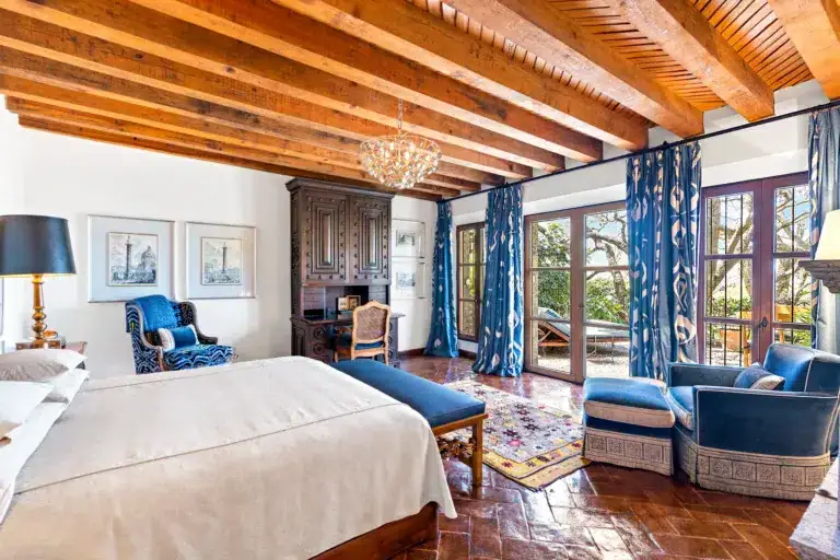 Living room with exposed wooden beam ceiling, crystal chandelier, and dark antique cabinet beside tall blue-curtained windows, San Miguel de Allende