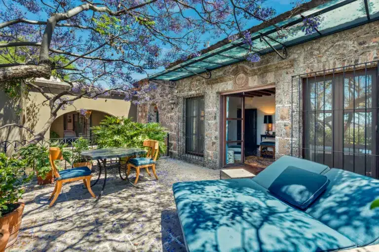 Stone patio courtyard with blue-cushioned lounge chairs, wooden chairs around a round table, and a jacaranda tree overhead with purple blossoms, San Miguel de Allende