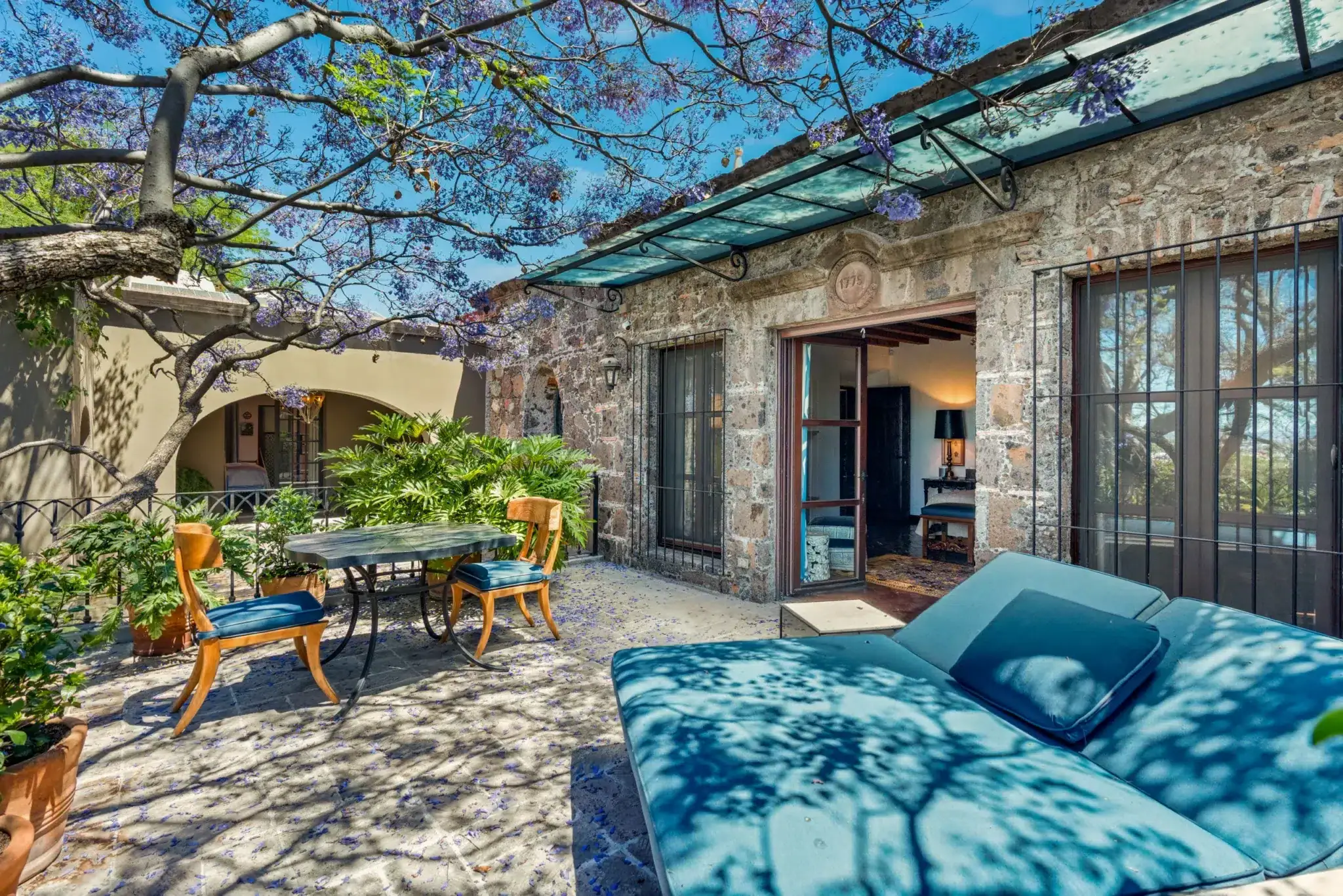 Stone patio courtyard with blue-cushioned lounge chairs, wooden chairs around a round table, and a jacaranda tree overhead with purple blossoms, San Miguel de Allende