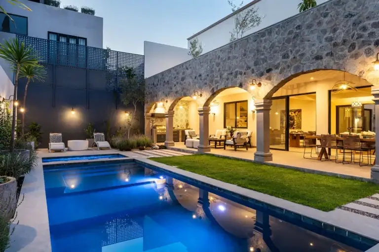 Modern backyard with a rectangular pool, stone arched colonnade, and an open-air dining area under warm lighting at dusk, San Miguel de Allende
