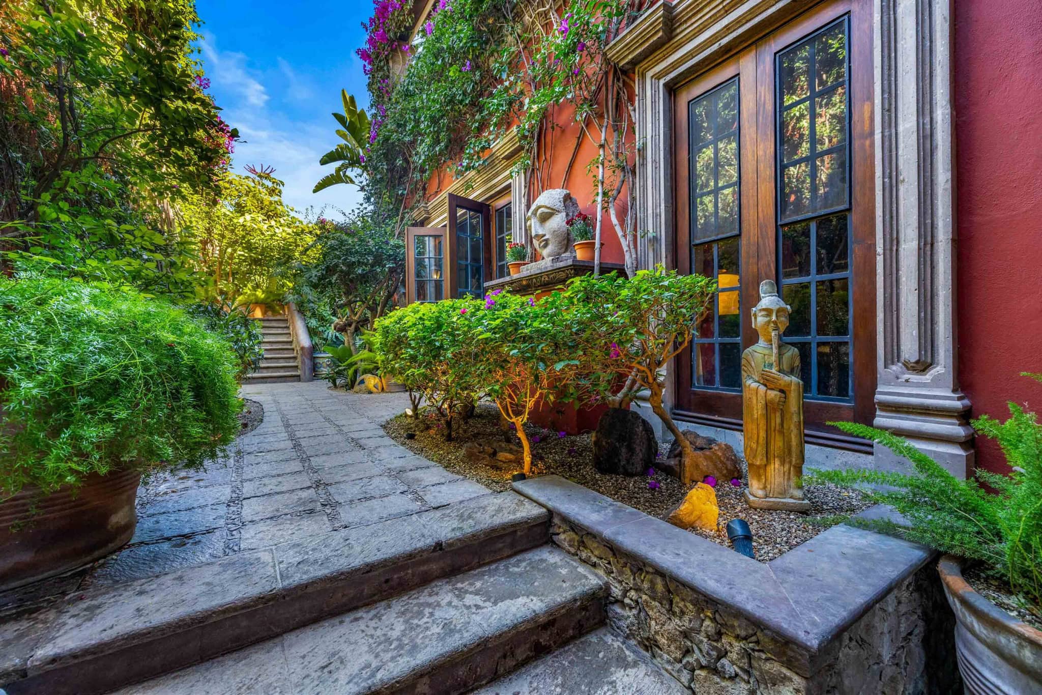 Stone steps lead up to a red-walled building with wooden-framed glass doors, surrounded by lush greenery and flowering plants, San Miguel de Allende