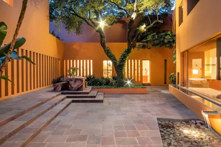 Orange-walled courtyard at dusk with a central tree and warm lighting, marble seating, and a glass-facing room on the right, San Miguel de Allende