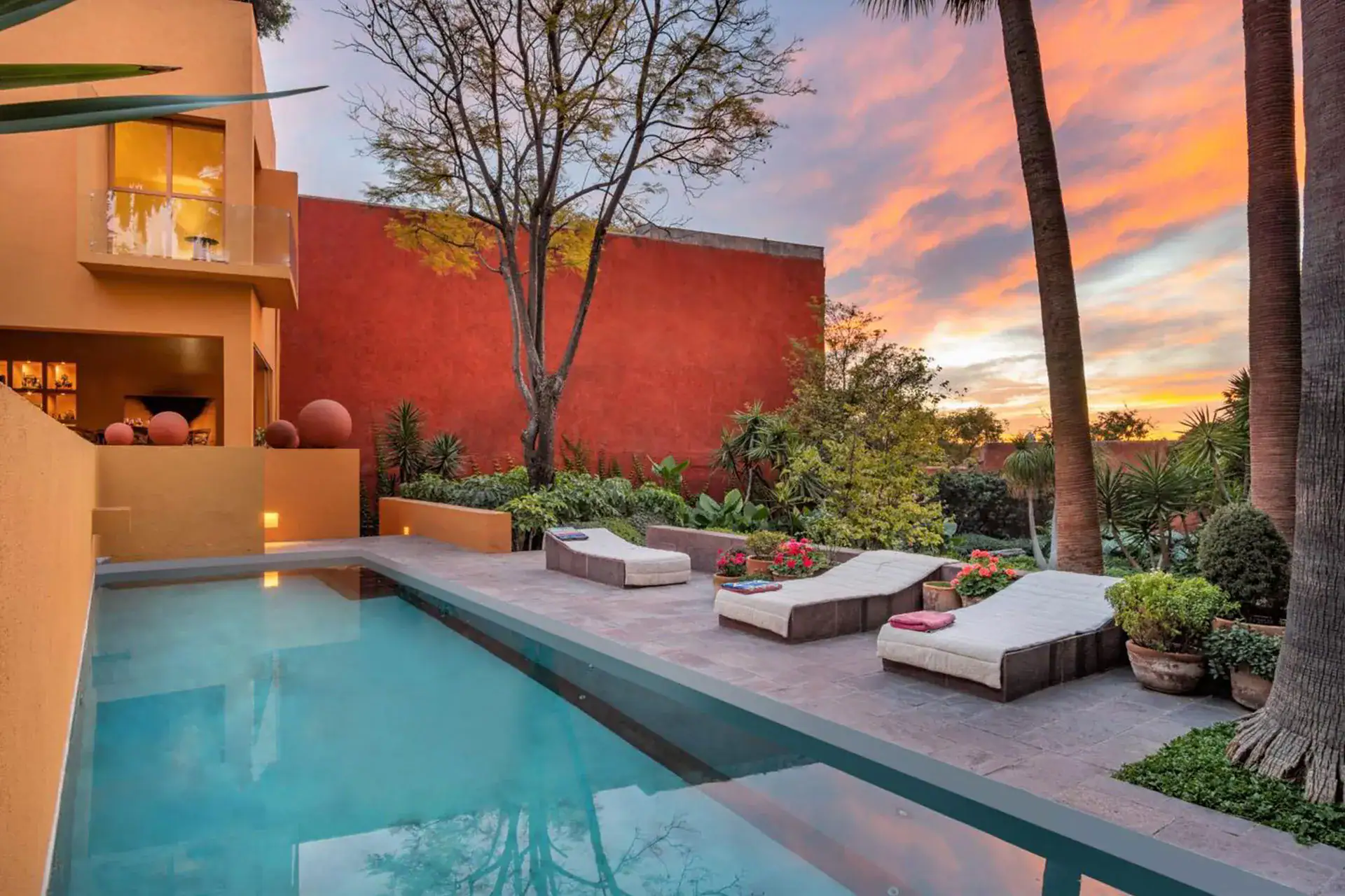 Modern villa courtyard with a rectangular pool, beige lounge chairs, orange walls, and palm trees at sunset, San Miguel de Allende