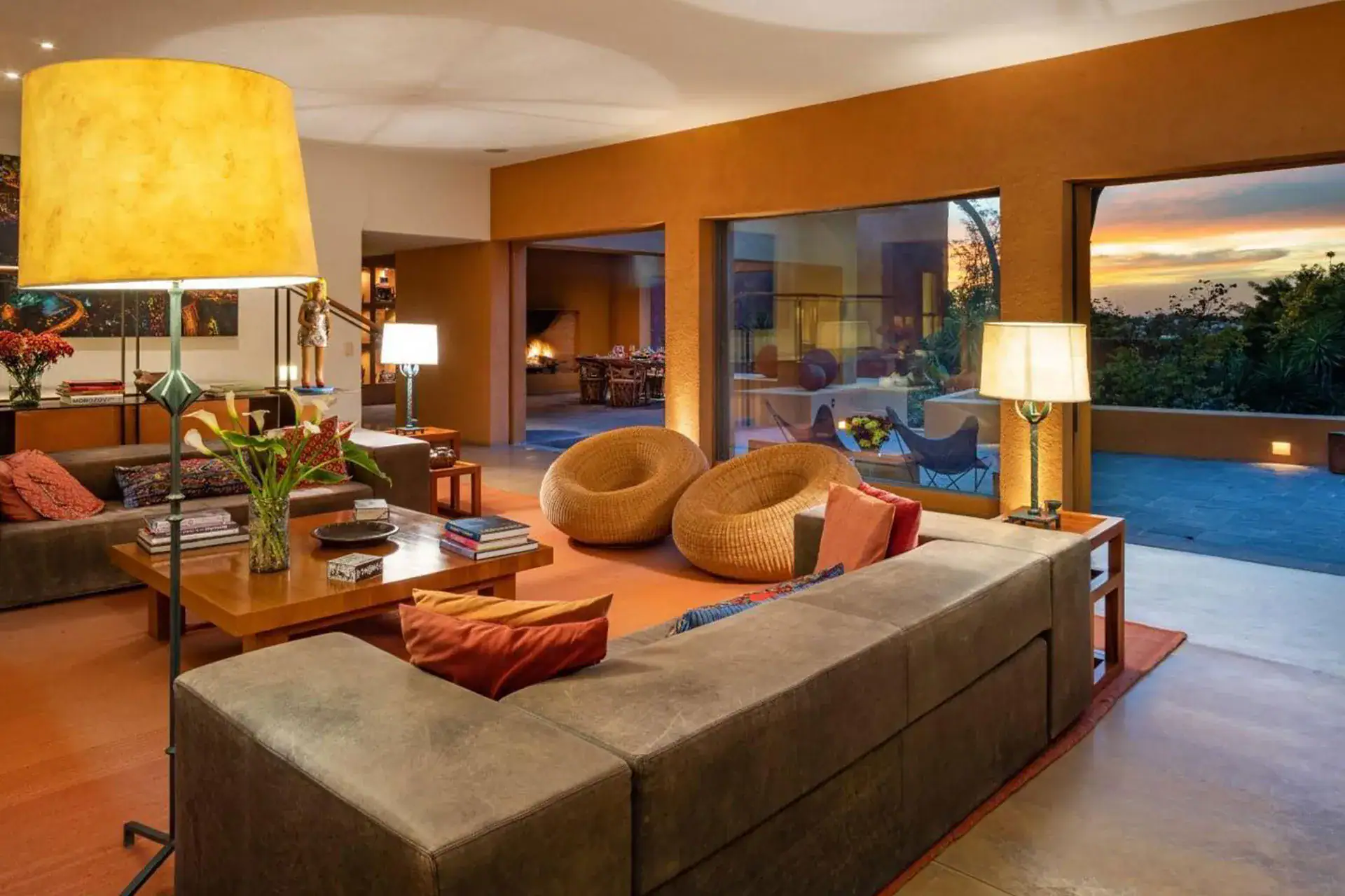 Spacious living room with a large sectional sofa, woven round chairs, and a coffee table stacked with books, illuminated by warm lamps; sliding doors open to a sunset-lit terrace, San Miguel de Allende
