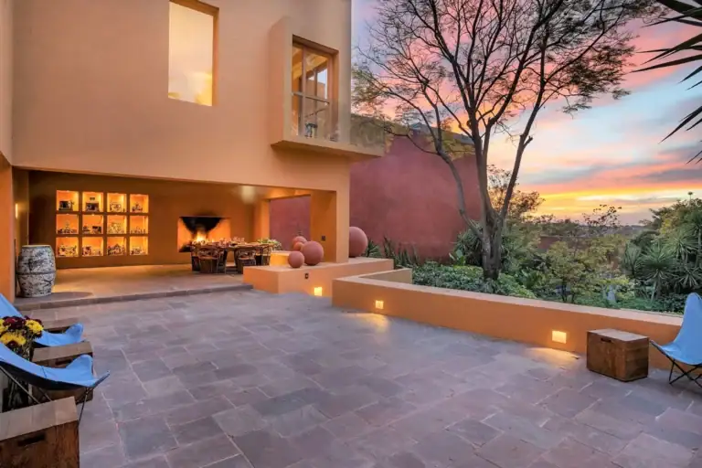 Terracotta-colored home exterior with a lit outdoor fireplace and dining table on a dusk-lit patio, San Miguel de Allende