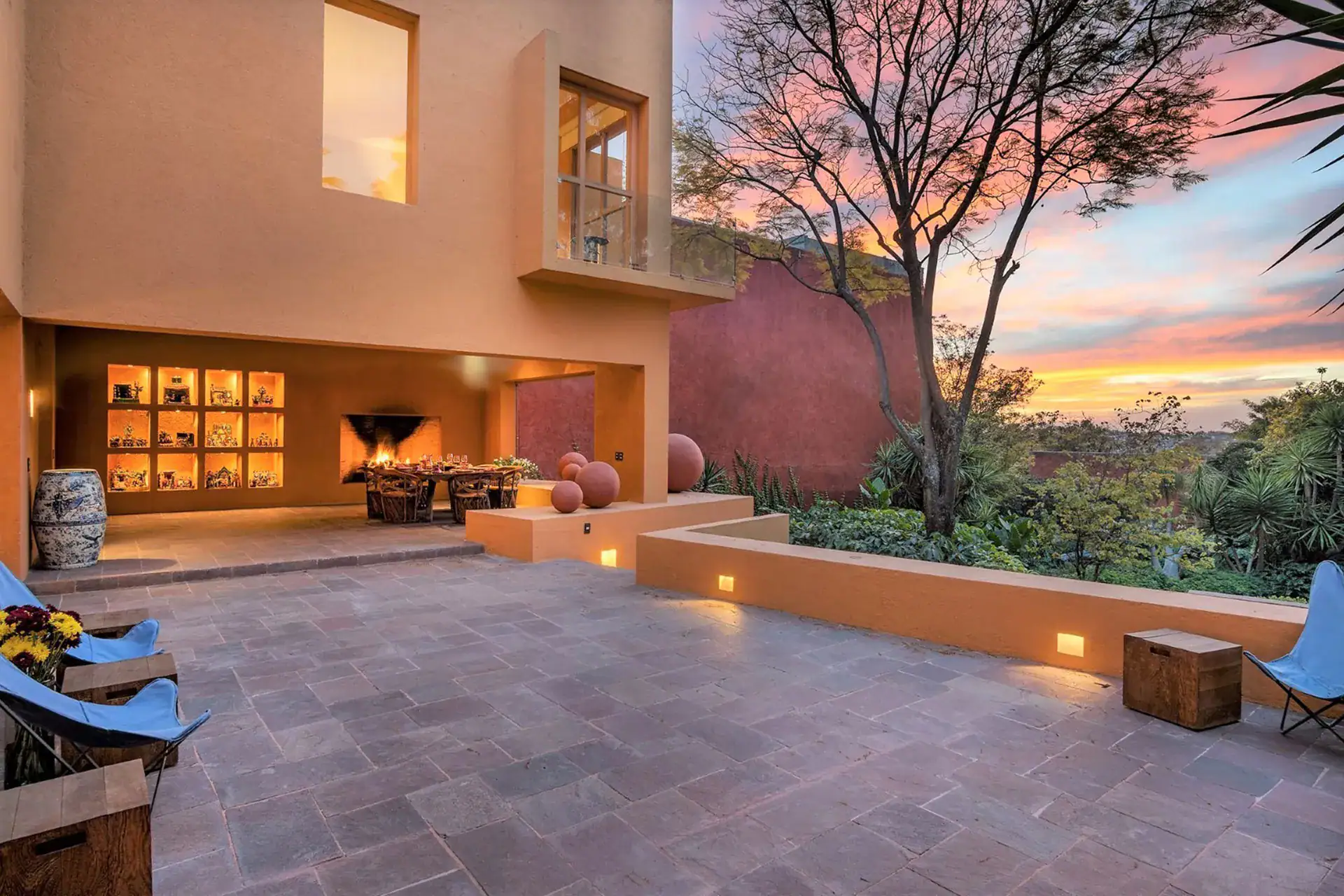 Terracotta-colored home exterior with a lit outdoor fireplace and dining table on a dusk-lit patio, San Miguel de Allende
