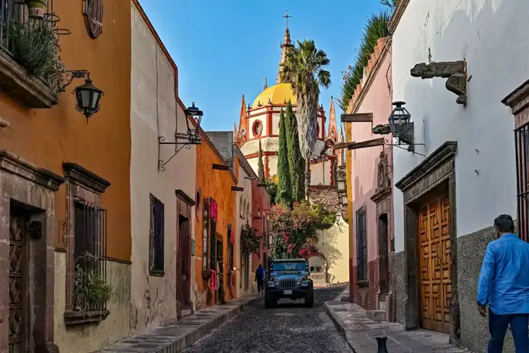 Colorful narrow street with pastel houses, a yellow-domed church at the end, a jeep approaching, and a man in blue on the right.