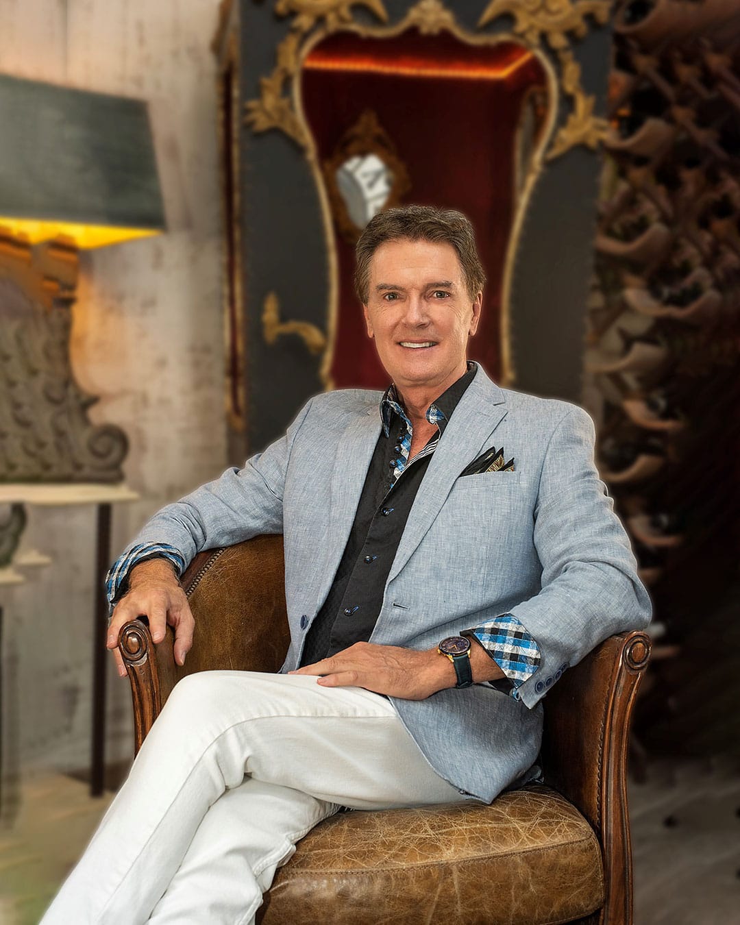 Man in a light blue blazer and white pants sits in a brown leather chair, smiling at the camera.