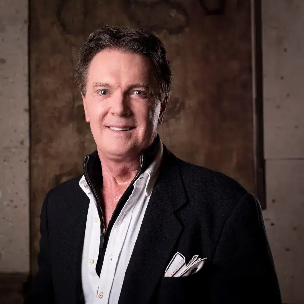 Portrait of a smiling man in a black blazer and white shirt, with a pocket square, against a rustic concrete backdrop.