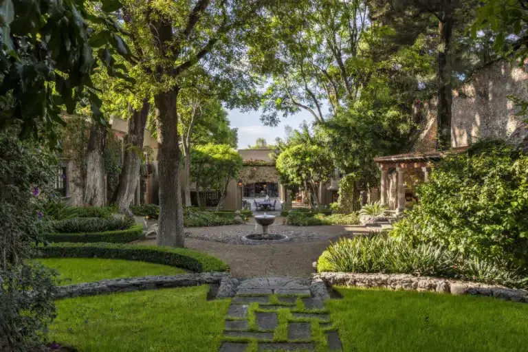 A tranquil stone courtyard framed by trees, with a central fountain and a meandering stone path surrounded by greenery, San Miguel de Allende