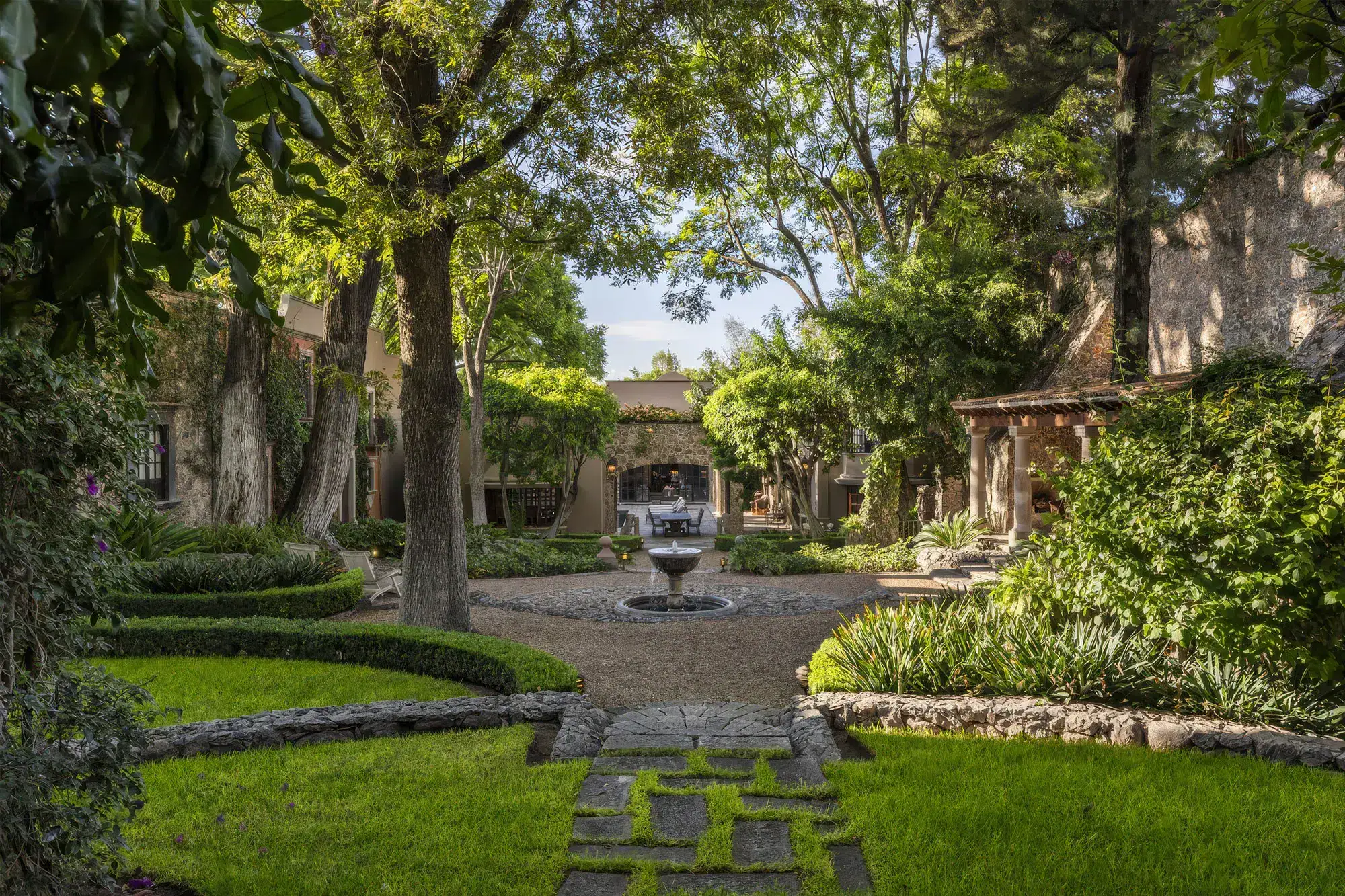 A tranquil stone courtyard framed by trees, with a central fountain and a meandering stone path surrounded by greenery, San Miguel de Allende