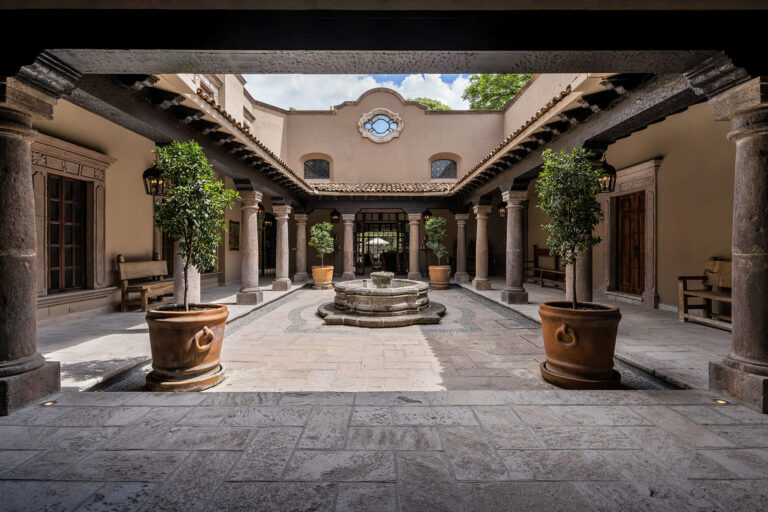 Open-air courtyard with stone columns, arches, and a central circular fountain, flanked by potted trees on a tiled floor, San Miguel de Allende