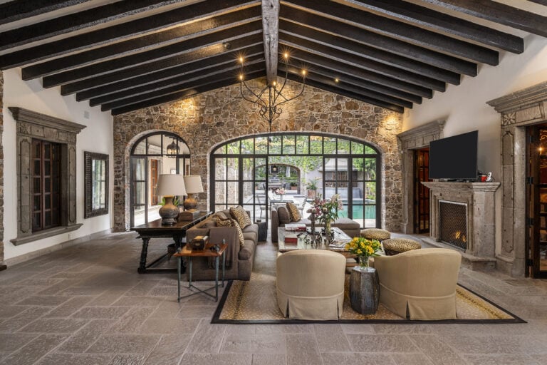 Rustic living room with dark wooden ceiling beams, stone walls, and a wrought-iron chandelier above a plush seating area, San Miguel de Allende