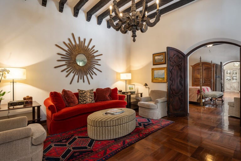 Living room with a red velvet sofa and patterned cushions, sunburst mirror on the wall, and a dark chandelier overhead, San Miguel de Allende