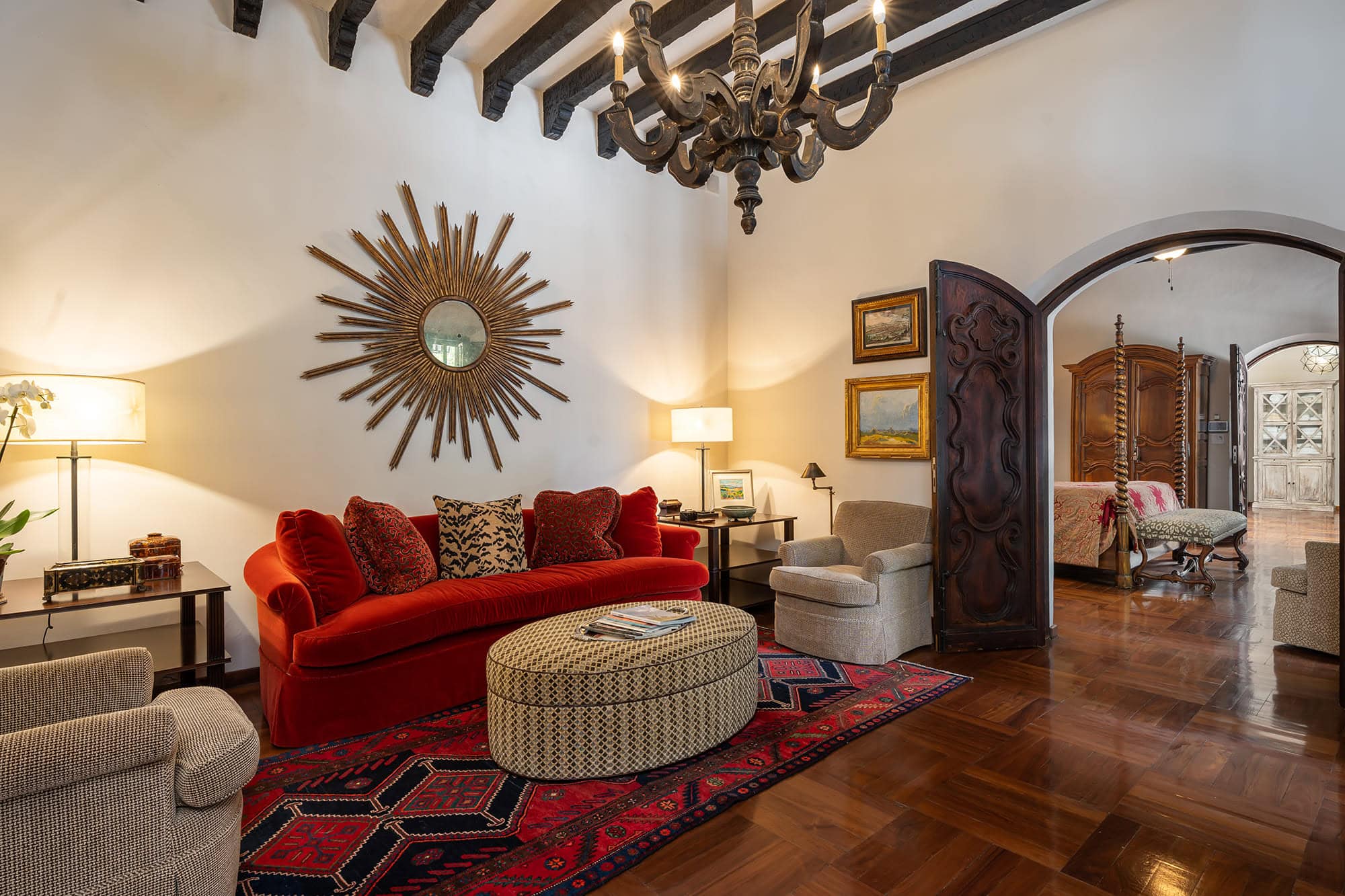 Living room with a red velvet sofa and patterned cushions, sunburst mirror on the wall, and a dark chandelier overhead, San Miguel de Allende
