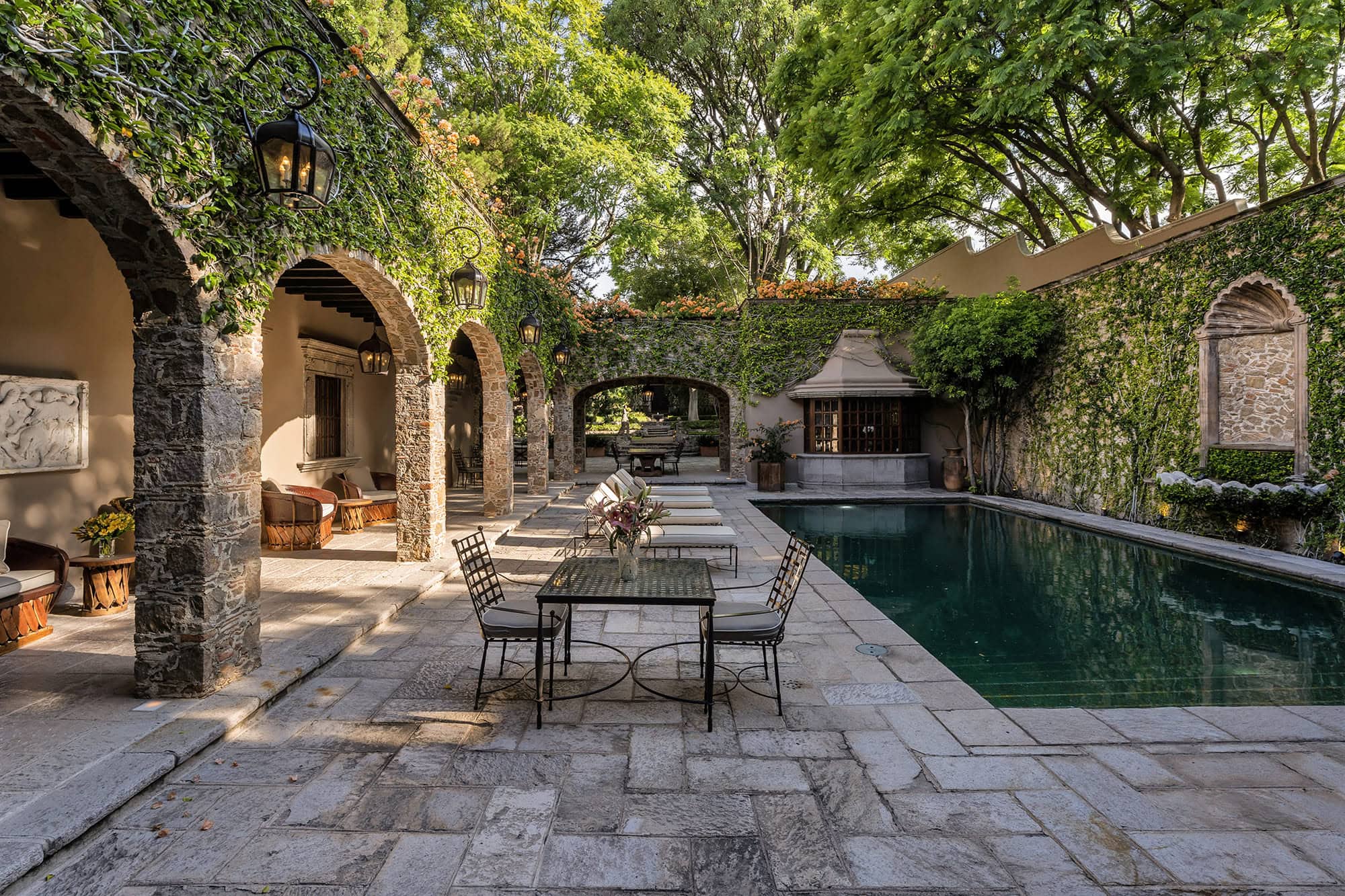 Stone courtyard with ivy-covered arches, lanterns, and a rectangular pool surrounded by patio seating and lush trees, San Miguel de Allende