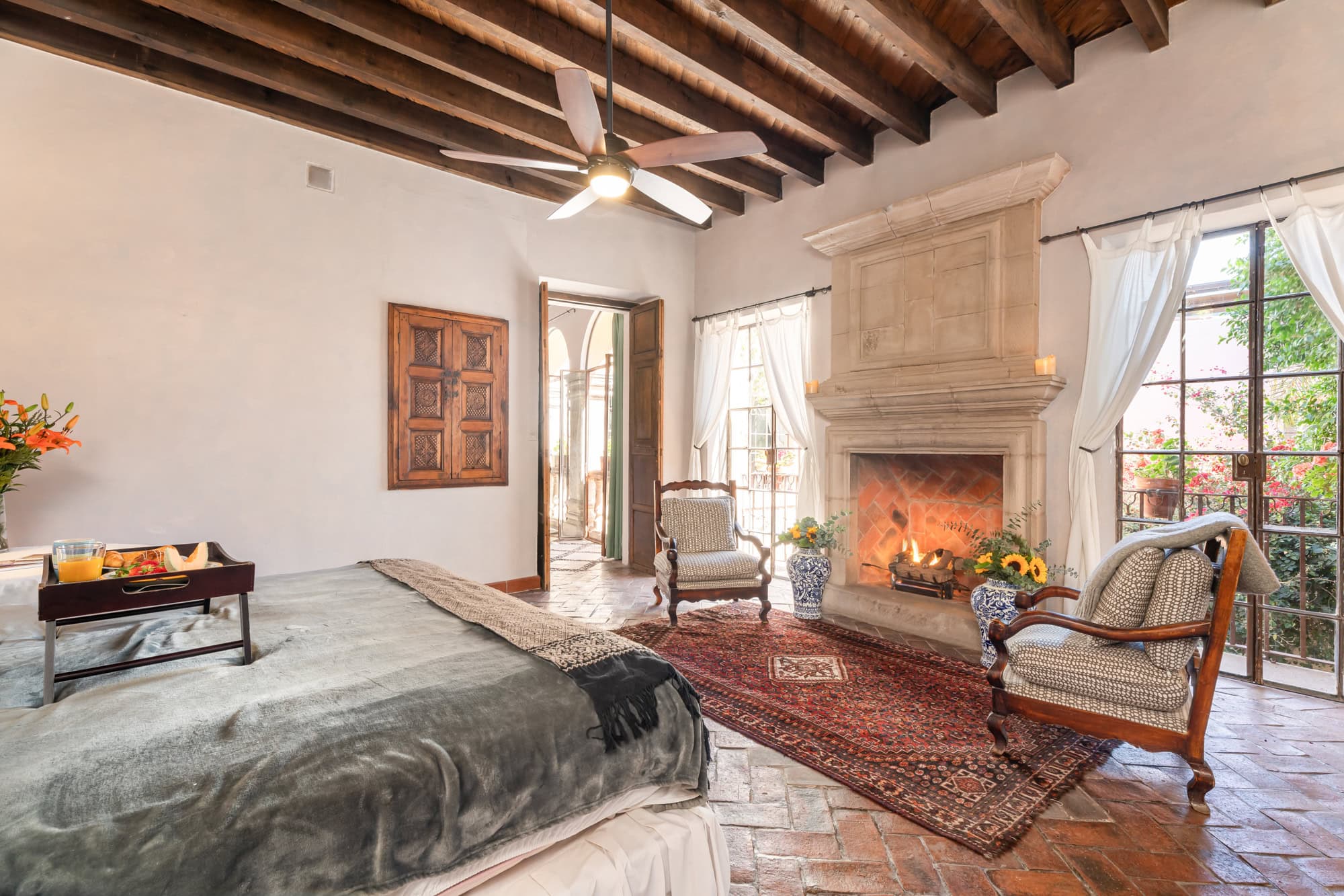 Sunlit living room with a stone fireplace, white walls, and large windows with white drapes showing greenery outside, San Miguel de Allende