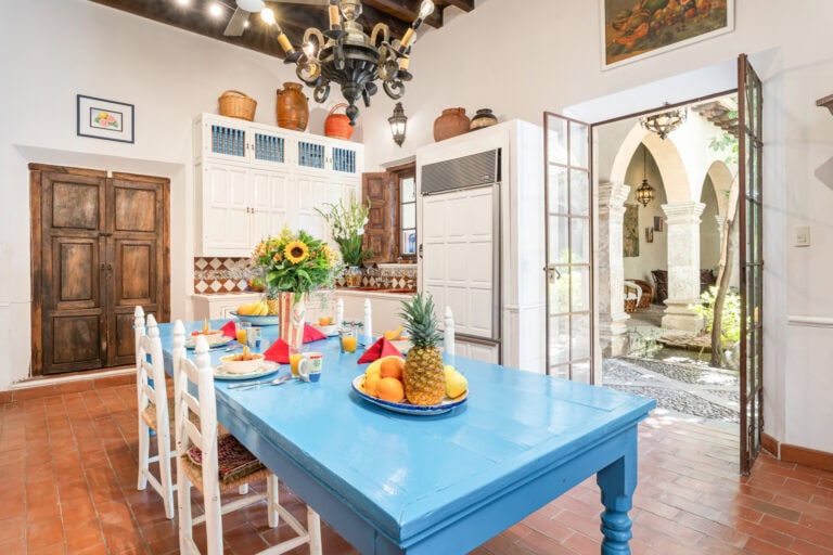 Bright dining area with a blue table set for a meal, white chairs, and a fruit centerpiece in a sunlit kitchen, San Miguel de Allende