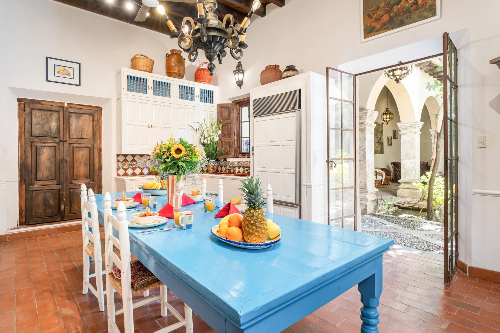 Bright dining area with a blue table set for a meal, white chairs, and a fruit centerpiece in a sunlit kitchen, San Miguel de Allende