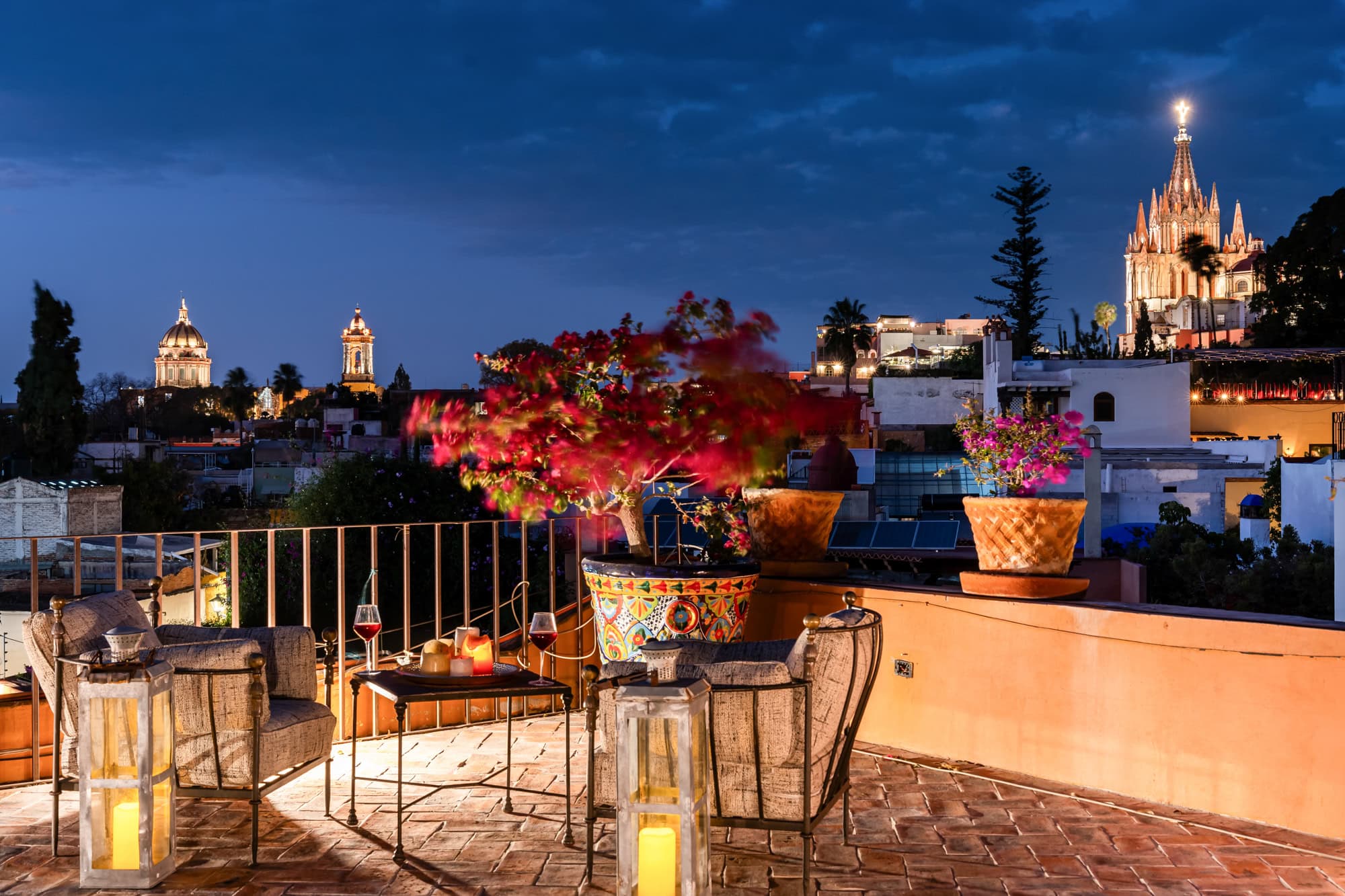 Rooftop terrace at dusk with lanterns, wicker seating, and colorful potted plants; a distant illuminated cathedral and city skyline in the background, San Miguel de Allende