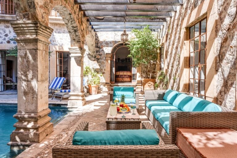 Sunny stone courtyard with arches, wicker lounge seating and turquoise cushions beside a pool, San Miguel de Allende