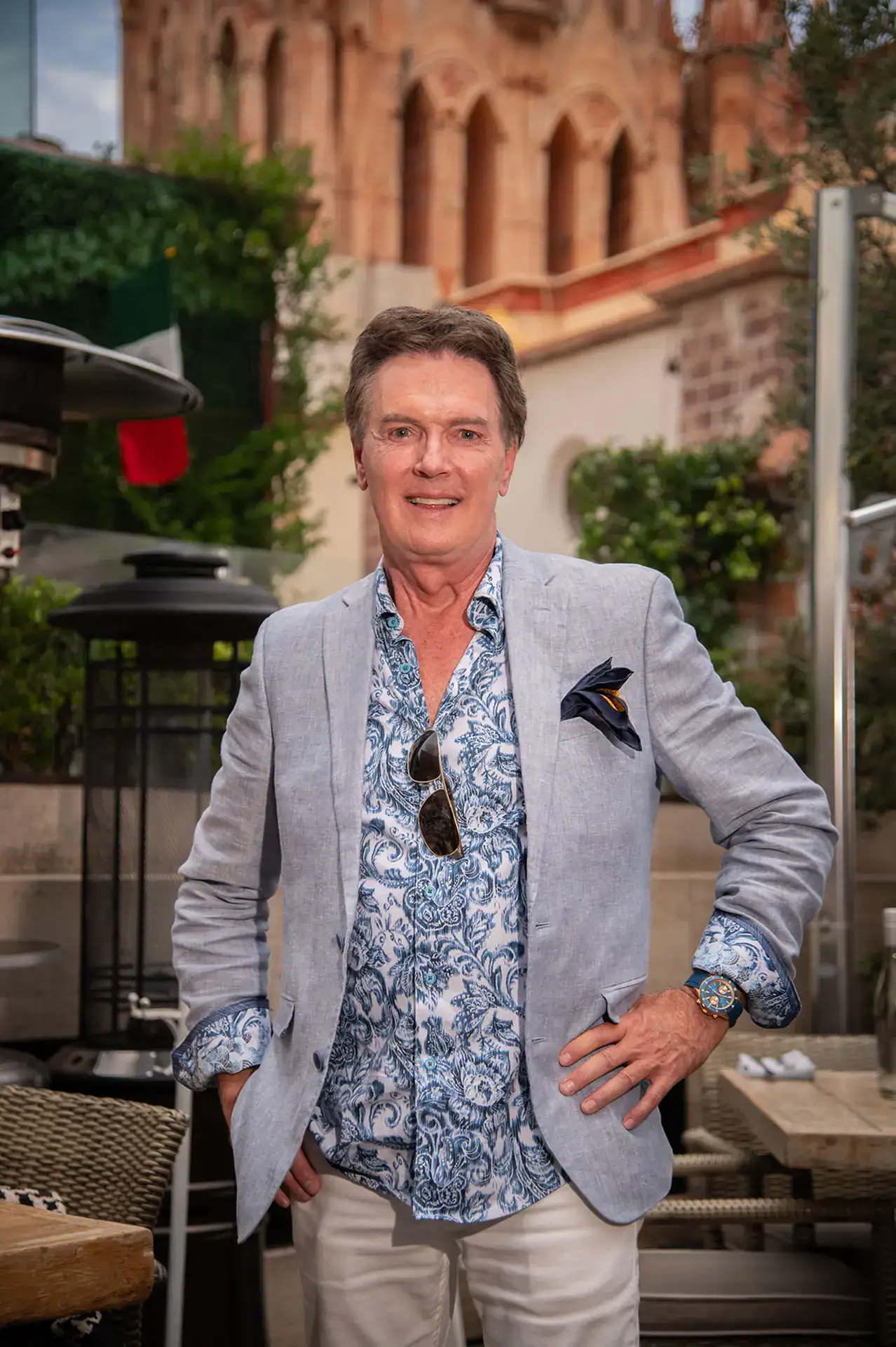 Smiling man in a light blue blazer and blue floral shirt posing outdoors at a cafe, sunglasses tucked in his shirt pocket.