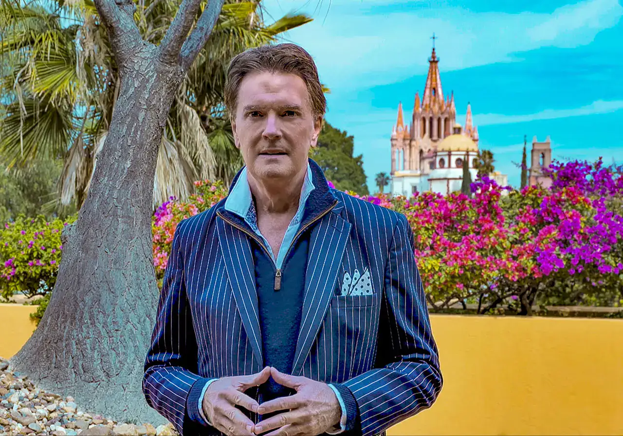 Man in a blue pinstripe blazer stands outdoors with colorful flowers and a church spire in the background.
