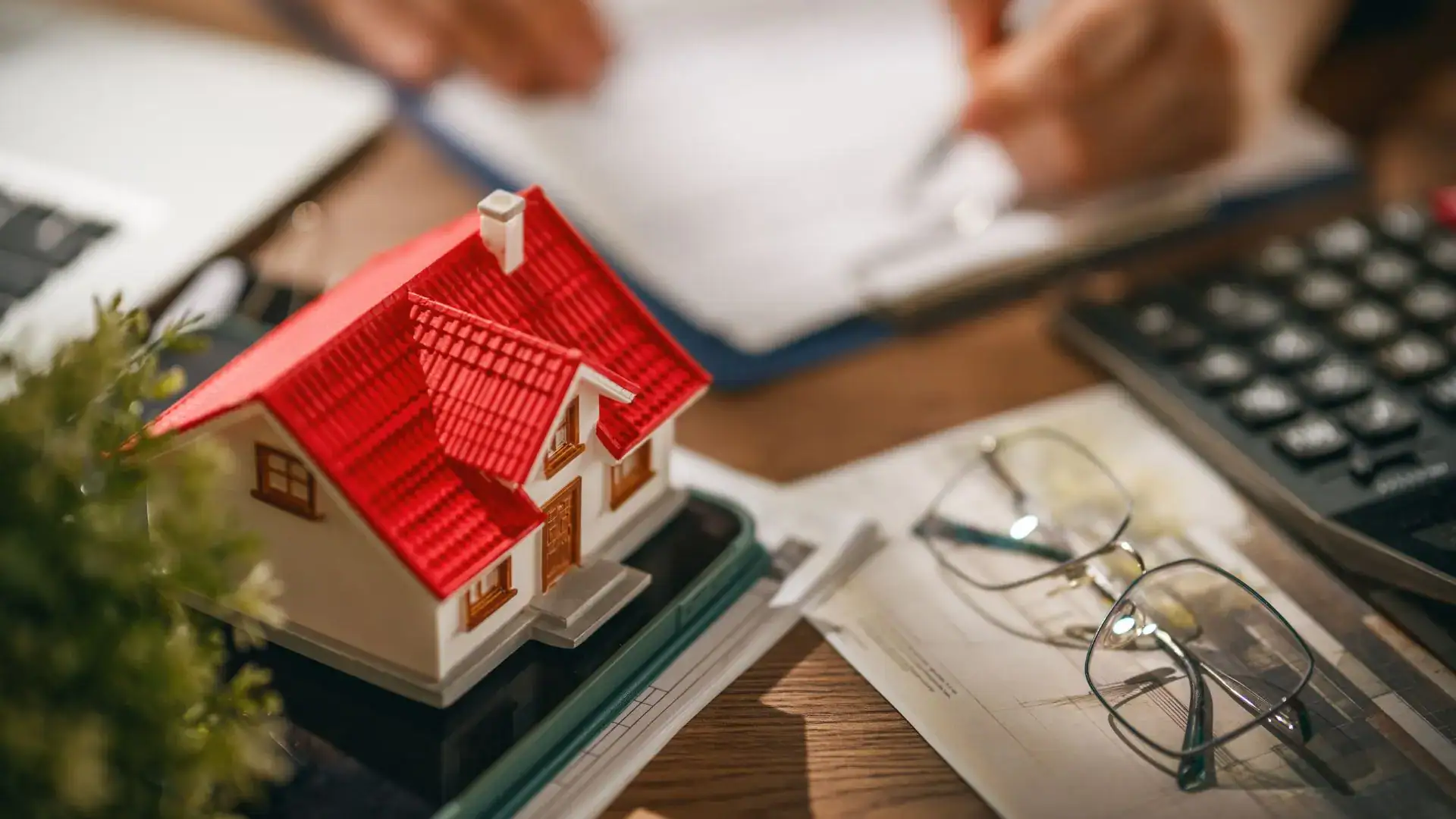 Model red-roofed house on a desk with papers, glasses, and a calculator, suggesting real estate planning.