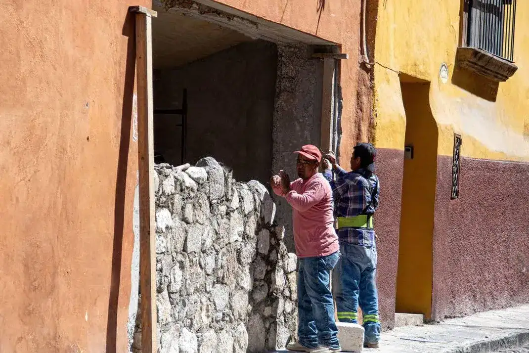 Two workers lay stones to build a low stone wall along a sunlit colorful street with orange and yellow walls.