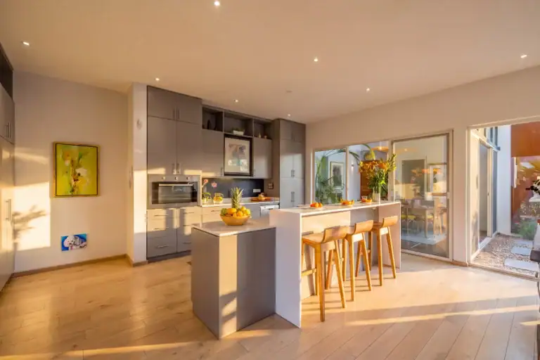 Modern kitchen with grey cabinets, central island, and wooden bar stools opening to a sunny patio area, San Miguel de Allende
