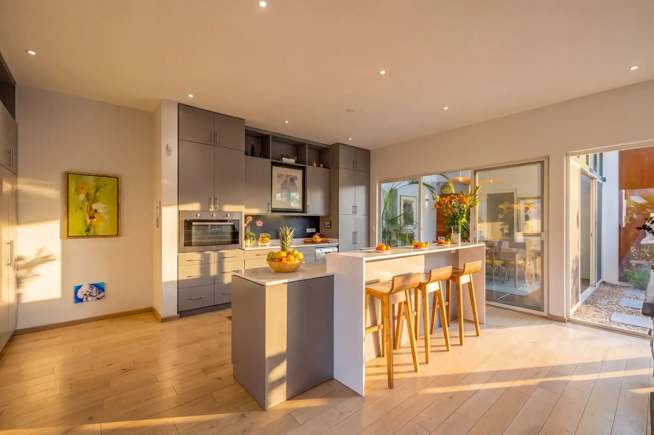 Modern kitchen with grey cabinets, central island, and wooden bar stools opening to a sunny patio area, San Miguel de Allende