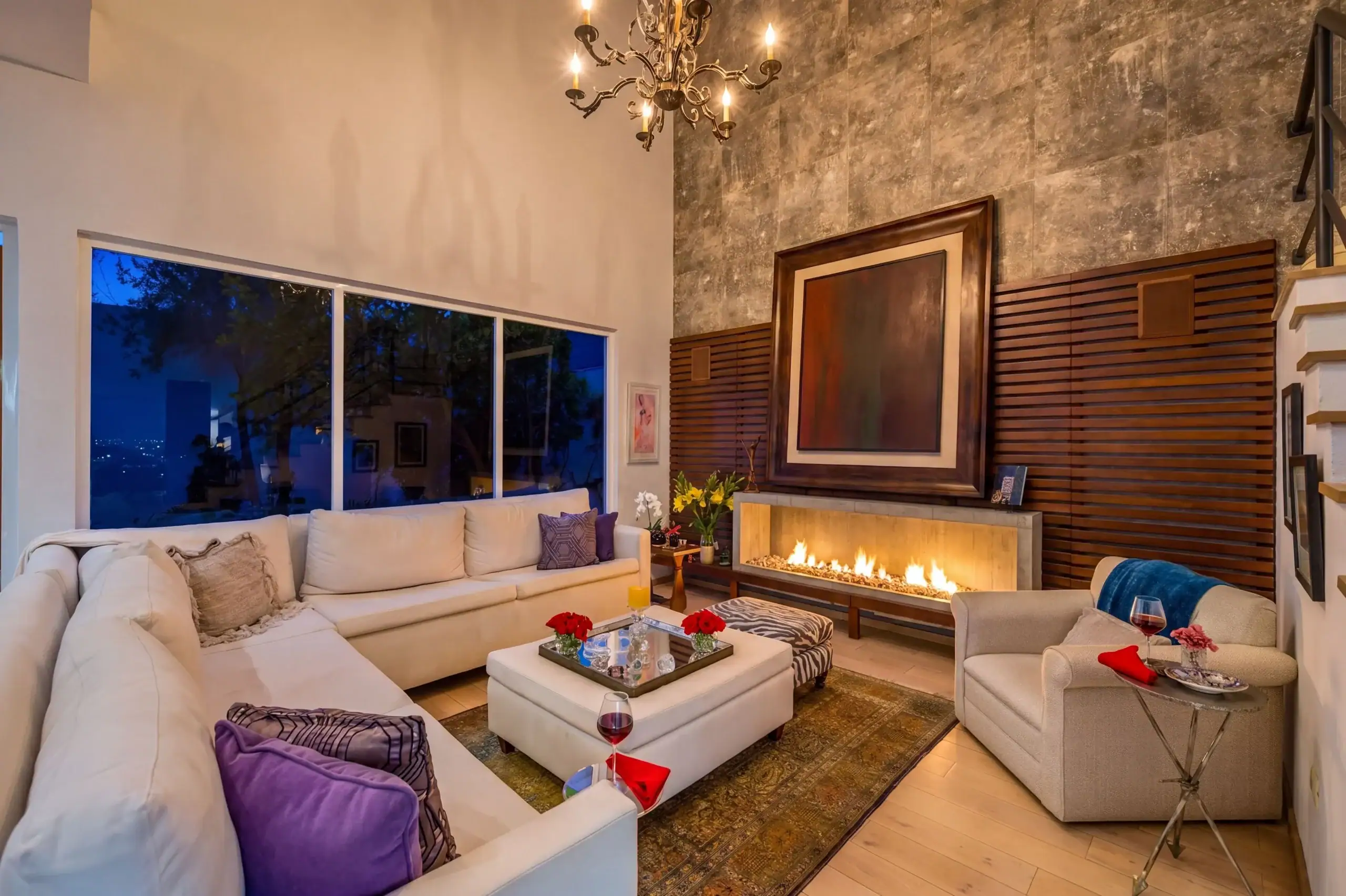 Cozy modern living room with a cream sectional, purple cushions, and a glass coffee table by a lit fireplace at dusk, San Miguel de Allende